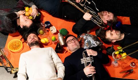 5 Dartellende liegen auf orangener Decke ungeben von Obst