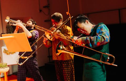 drei musizierende, zwei spielen im hintergrund ihre blasinstrumente, im vordergrund versucht ein dritter seine posaune zu richten 